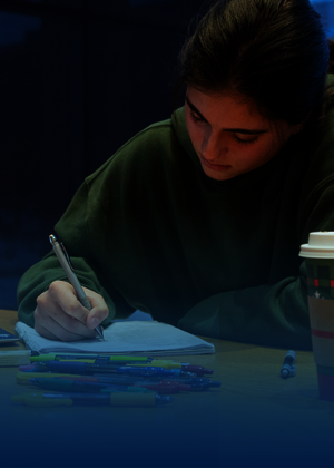 Student studying at a desk with a laptop and coffee cup, in dim lighting.
