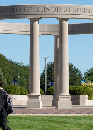 Circular stone structure with tall columns, people walking in front on a sunny day.