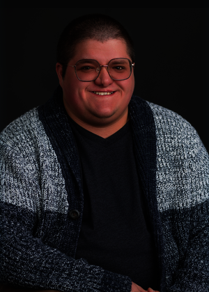 Smiling person in glasses and a blue-gray sweater against a dark background.