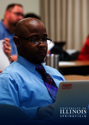 Man in a blue shirt and glasses works on a laptop in a classroom setting.