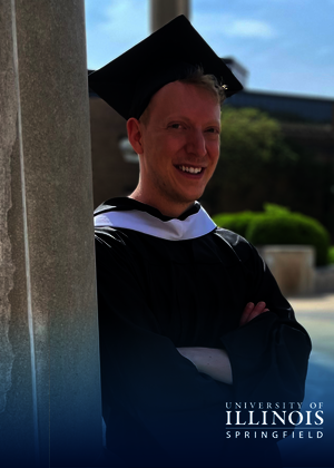 Graduate smiling in cap and gown, standing outside near a pillar.