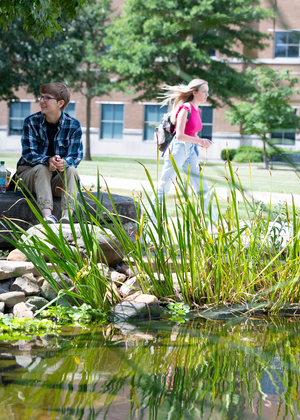 Students by a pond on a sunny day, university building in background.