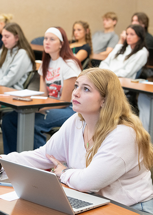 Students attentively listening in a classroom with laptops.