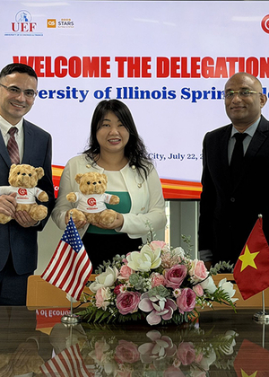 Three people smiling, holding teddy bears, with flags and flowers on a table.