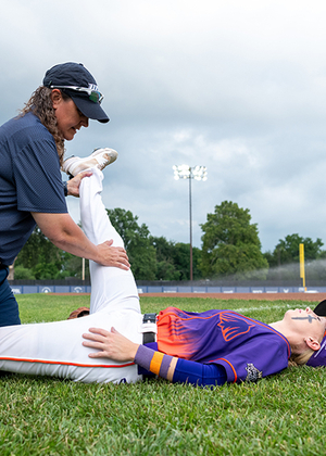 Baseball coach helps player stretch leg on field, cloudy sky.