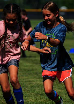 Two girls running on a soccer field, smiling and competing closely.