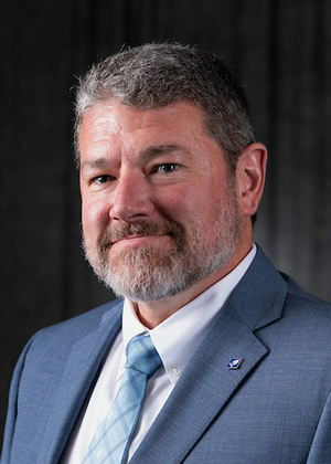 Man with a beard in a blue suit and tie against a dark background.