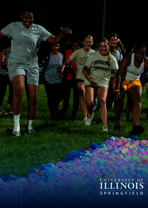 Students running towards colorful balls on a grass field at night.