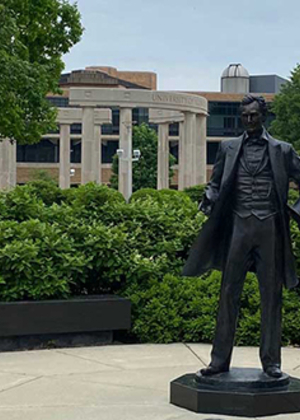 Statue of Abraham Lincoln standing in a garden area with the Colonnade and campus buildings behind.