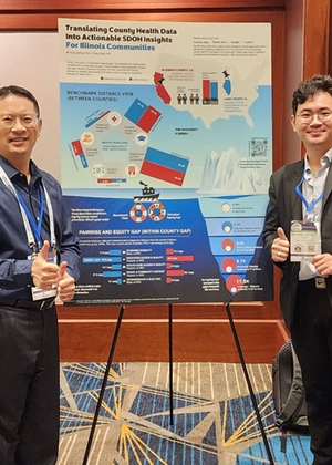 Two men smiling and giving thumbs up, standing beside a colorful conference poster on a stand.