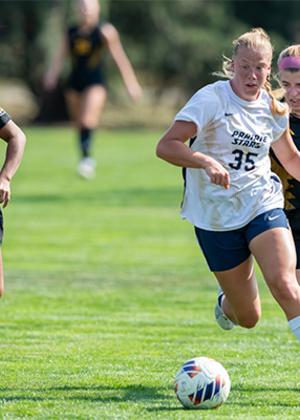 UIS soccer players in action on a green field, one leading with the ball.
