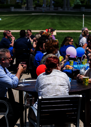 Outdoor gathering with people seated at tables, balloons, and sunny weather.