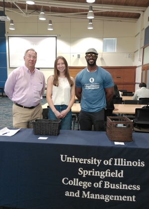 Three people standing behind a university information table indoors.