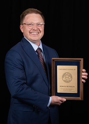 Man in suit holding a wooden plaque, smiling against a black background.
