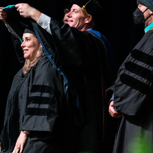 In a ceremonial gesture, the associate dean Travis Bland presents a graduation cap to a beaming graduate, marking the completion of their studies.