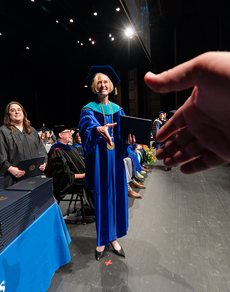 A university official in academic regalia extends a diploma and handshake to a graduate during a commencement ceremony.