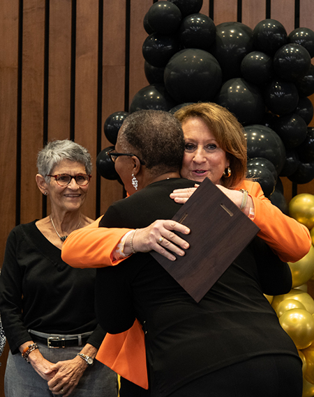 A person receives an award and a hug during the Good as Gold Volunteer Awards ceremony, with others smiling nearby.