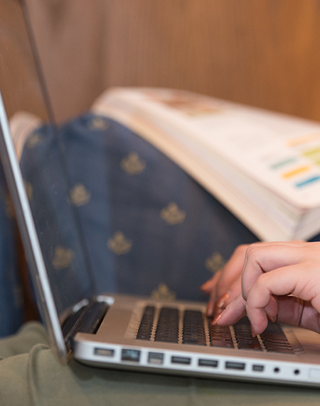 Close-up of a person typing on a laptop with an open textbook in the background.
