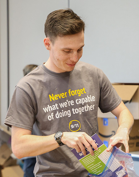A person packing a colorful bag with supplies in a room filled with boxes. Their shirt reads, "Never forget what we're capable of doing together." 