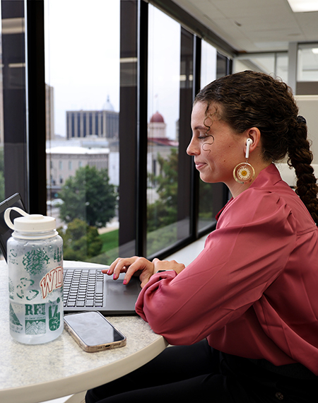 Young woman works on a laptop at a high-rise office window with earbuds, water bottle, and phone nearby.