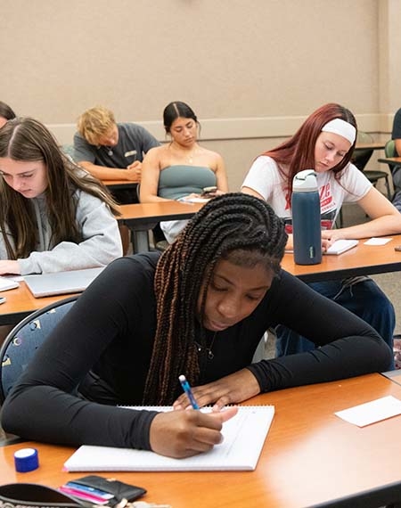 Students sit at desks in a classroom, writing notes and focusing on their work.