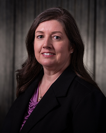 Smiling woman with dark hair in a dark room, wearing a purple shirt.