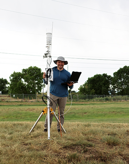 Man with tablet stands by weather station in grassy field.