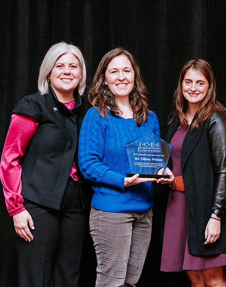 Three women smiling, one holding an award in a blue sweater. Black background.