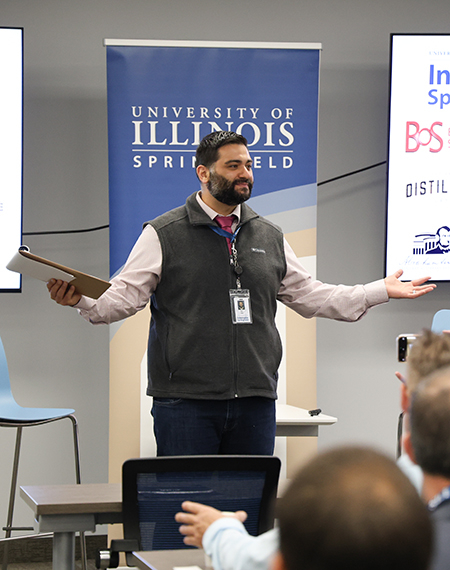 Man speaking at a presentation, University of Illinois Springfield banner behind him.
