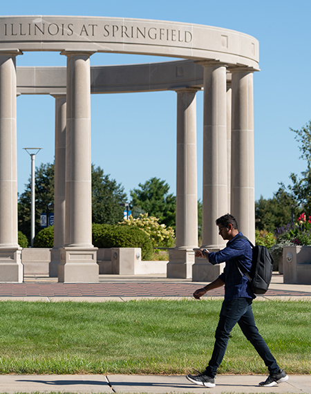 Man walks past stone pillars in park setting under clear blue sky.