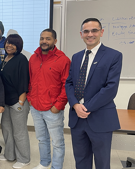 Group of people standing in a classroom, one in a suit, others in casual attire.