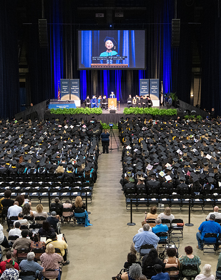 Graduation ceremony with speaker on stage, audience in caps and gowns.