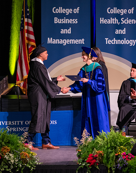 Graduate receives diploma on stage from faculty member in a blue robe.