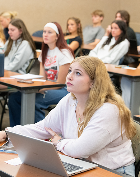 Students attentively listening in a classroom with laptops.