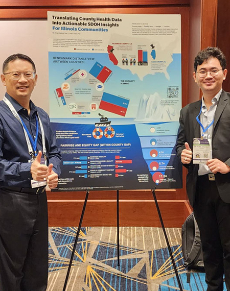 Two men smiling and giving thumbs up, standing beside a colorful conference poster on a stand.