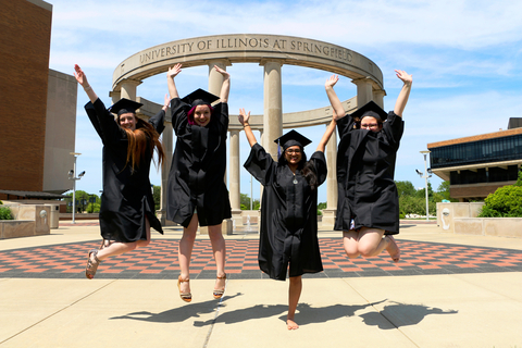 Four graduates jumping in front of the UIS colonnade
