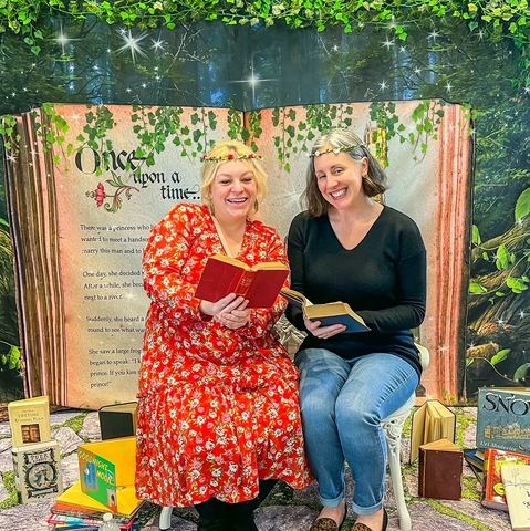 Two women sitting on a bench reading books with a backdrop of a book behind them.