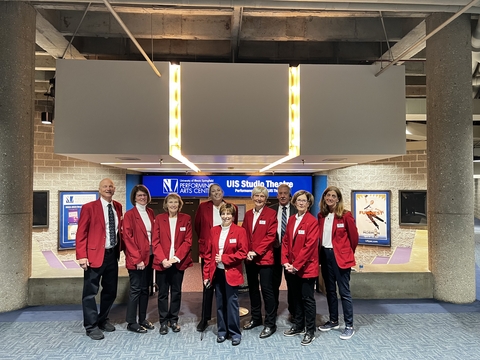 A group of volunteers wearing red coats and posing in front of the UIS Studio Theatre for a photo