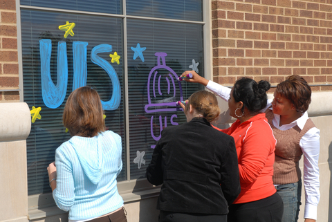 four people paint UIS themed images on a window on a building at UIS