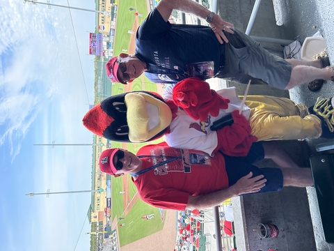 two people pose with a Redbird mascot in front of a baseball field