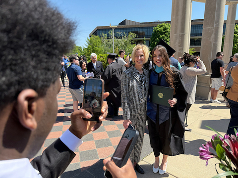 Chancellor Gooch poses with a graduate in front of the colonnade while someone takes their photo on a phone.