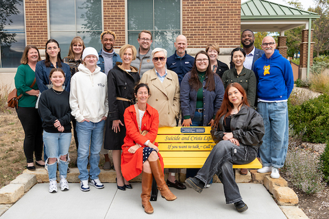 Campus community members sit on and stand around new yellow bench for suicide awareness
