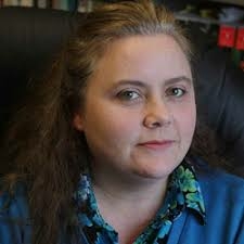 A woman with long brown hair, wearing a blue blouse, looks calmly at the viewer.