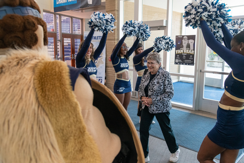 Cheerleaders and a mascot welcome a woman indoors.