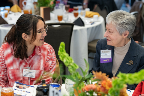 Two women smiling and talking at a table set with drinks and floral centerpiece.
