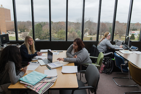 Four students studying at tables with papers everywhere in a room with large windows.