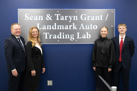 Four people in formal attire stand smiling in front of a blue wall with a metallic sign.