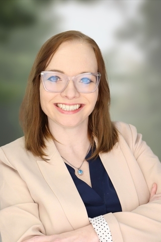 Smiling person in glasses and beige blazer, arms crossed.