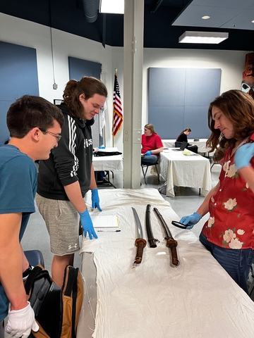 Three people in a room examine swords on a table, wearing blue gloves. An American flag and others in the background create a classroom atmosphere.