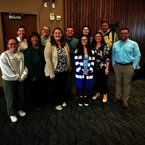 Eleven members of the strategic communication team smile in the Student Union Ballroom with CARE winner Ashley Earnst in the center.